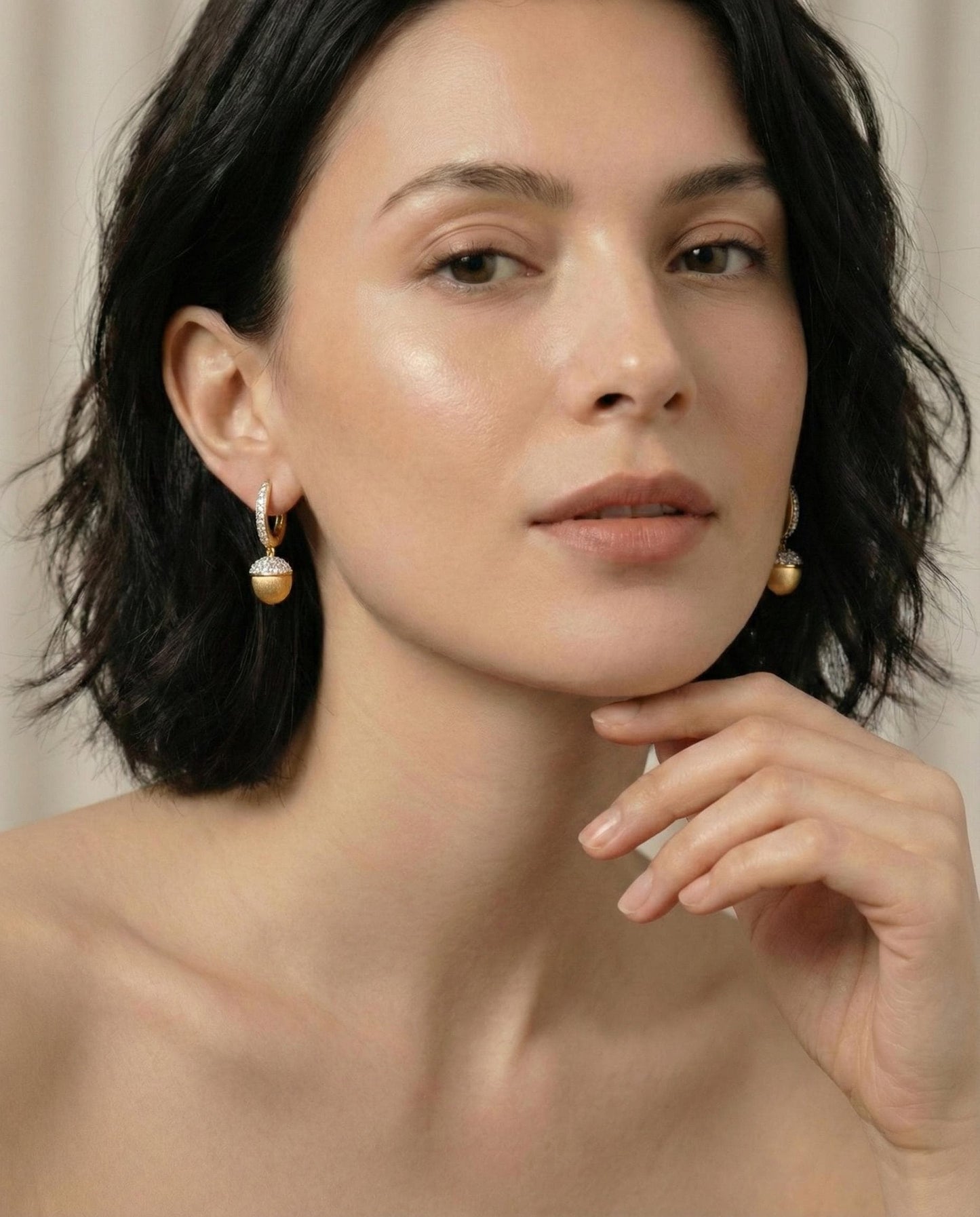 Woman with dark hair and earrings, wearing minimal makeup against a neutral background